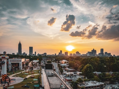 Sunset over Atlanta skyline, with clouds and vibrant colors, viewed from a rooftop with greenery and urban structures below.