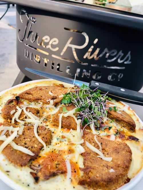 A plate of baked dish with toppings, in front of a Three Rivers Distilling Co. sign.