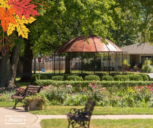 A gazebo surrounded by greenery and flowers, with benches nearby, set in a park during autumn.