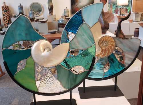 Two decorative glass sculptures resembling nautilus shells, featuring vibrant colors and intricate patterns, displayed in an art gallery.
