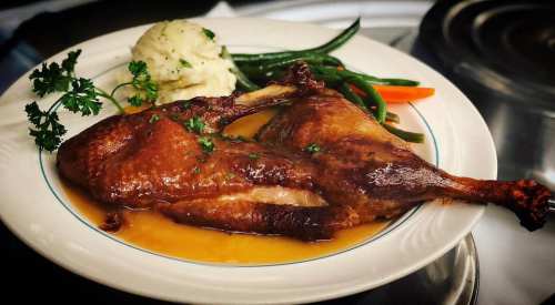 A plate of roasted duck with gravy, served alongside mashed potatoes and green beans, garnished with parsley.