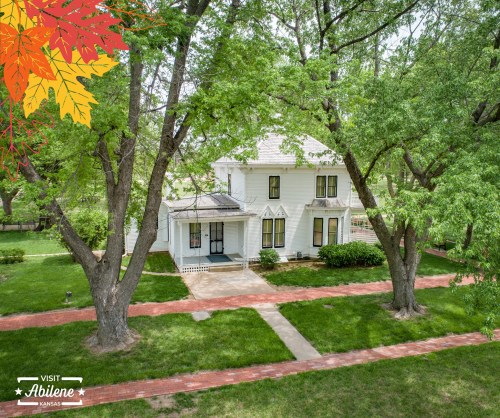 A charming white house surrounded by green trees and a red brick path, with a fall-themed graphic in the corner.