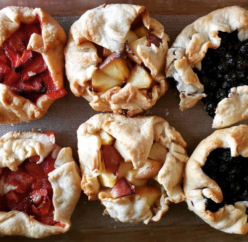 A tray of six rustic fruit galettes with various fillings: blueberry, apple, and strawberry.