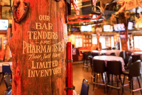 A rustic bar post with a humorous sign about bartenders being like pharmacists, set in a lively pub atmosphere.