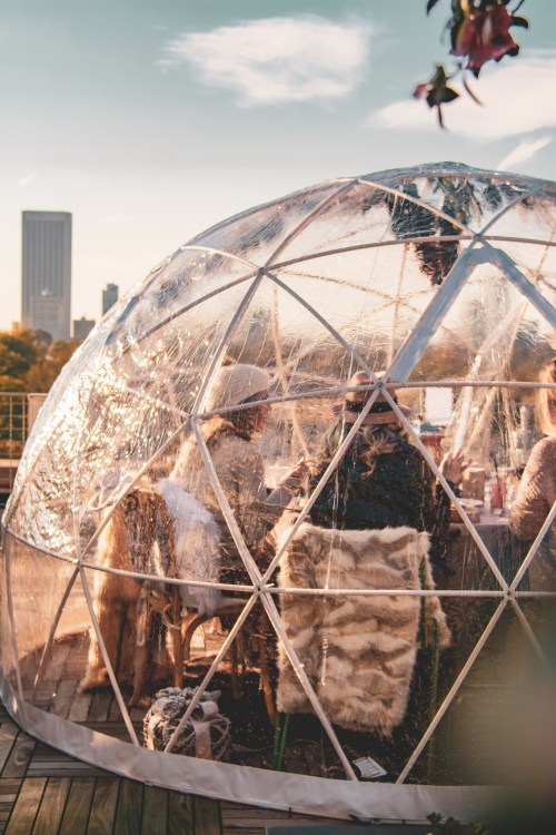 A transparent geodesic dome on a rooftop, with people dining inside against a city skyline backdrop.