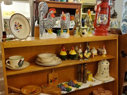 A wooden shelf displays various rooster figurines, dishes, and a vintage lantern in a cozy antique shop.