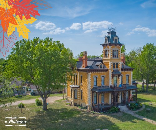 A historic yellow mansion surrounded by trees and grass, with a decorative roof and autumn leaves in the corner.