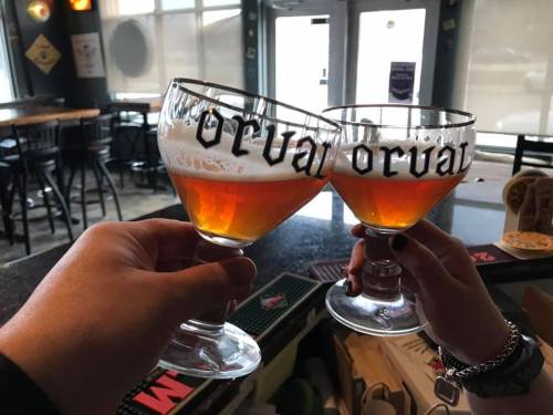 Two hands clink glasses of amber beer, each labeled "Orval," in a bar setting with empty tables in the background.
