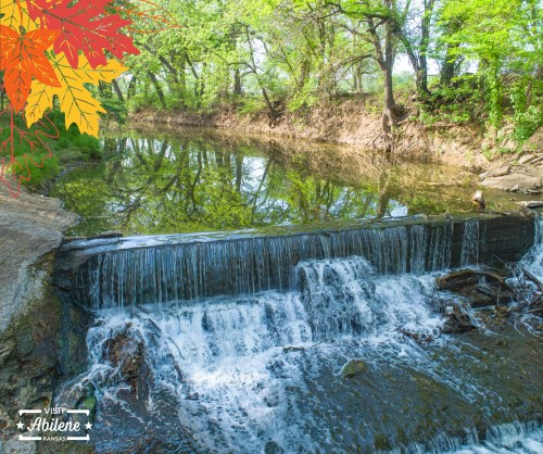 A serene waterfall flows into a calm river, surrounded by lush green trees and autumn leaves in Abilene, Kansas.