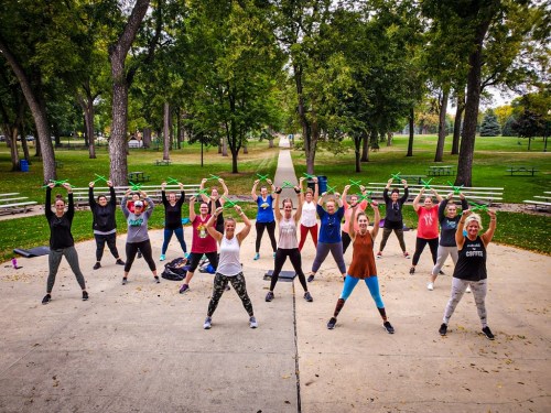 A group of women exercising outdoors in a park, holding green exercise bands and smiling.