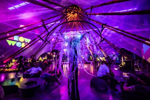 A vibrant indoor space with colorful lights, featuring a large tent structure and people socializing around low tables.