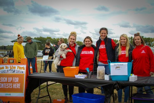 A group of five women in red shirts stand behind a table at an outdoor event, smiling and holding a small dog.