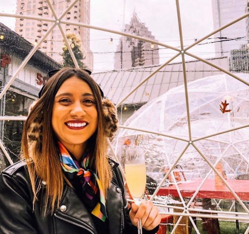 A smiling woman in a black jacket and colorful scarf holds a drink, with a transparent dome structure in the background.