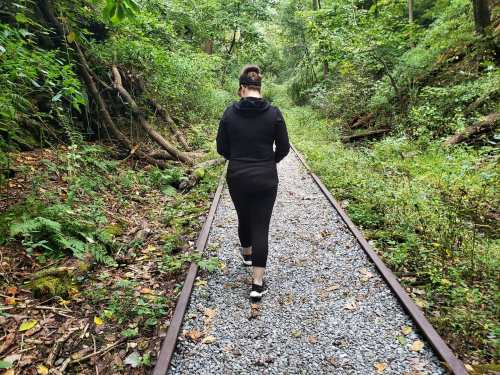 A person walks along a gravel path beside overgrown train tracks in a lush, green forest.
