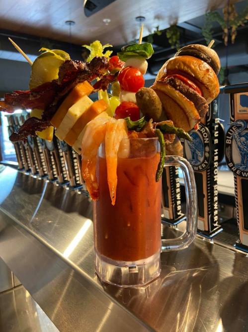A colorful, garnished cocktail with shrimp, bacon, cheese, and vegetables, served in a large glass on a bar counter.