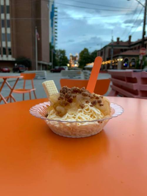 A dessert bowl with noodles, topped with crushed cookies and a wafer, sits on an orange table in an outdoor setting.