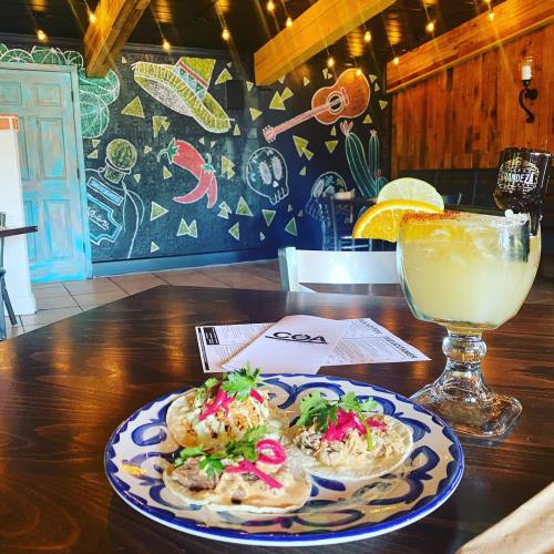 A colorful plate of tacos with toppings and a margarita on a wooden table, with a vibrant mural in the background.