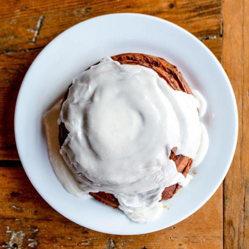 A cinnamon roll topped with creamy white icing on a white plate, resting on a wooden table.