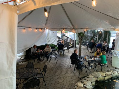 Outdoor dining area under a tent, with people seated at tables, string lights, and a pond in the background.