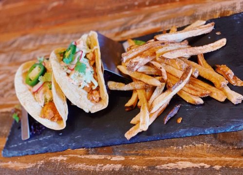 Two tacos filled with chicken, jalapeños, and toppings, served with a side of crispy French fries on a slate platter.