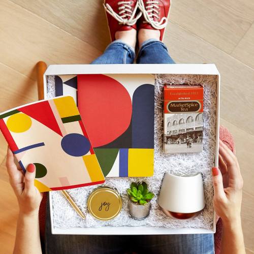 A person holds a gift box containing a notebook, a plant, a candle, and colorful stationery on a wooden surface.