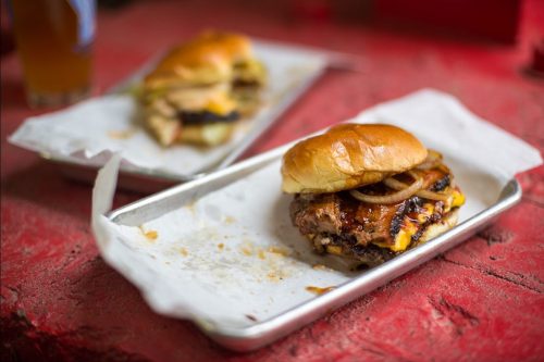 A close-up of a juicy burger with cheese and onions on a tray, with another sandwich in the background.