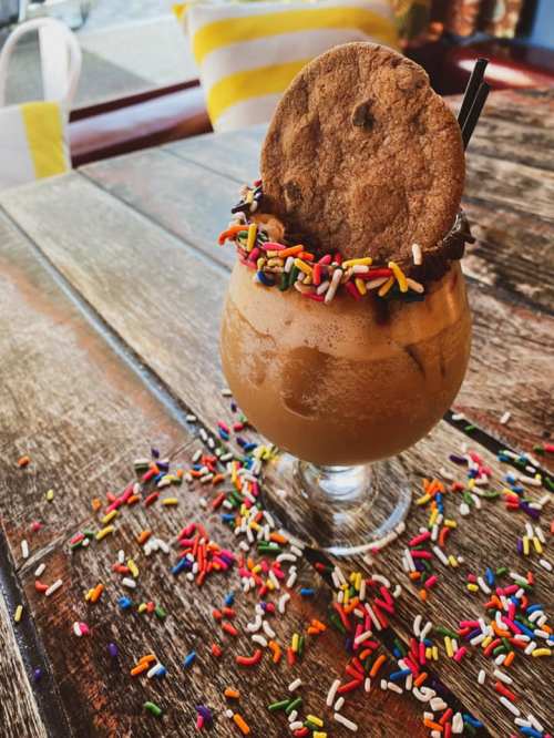 A frosty dessert drink topped with a cookie and colorful sprinkles, surrounded by more sprinkles on a wooden table.