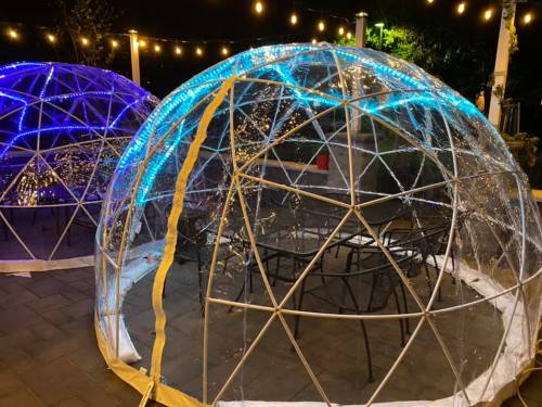 Two illuminated transparent domes with string lights, set up outdoors at night, surrounded by chairs and tables.