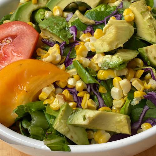 A colorful salad with corn, avocado, red and yellow tomatoes, and mixed greens in a white bowl.