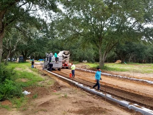 Construction workers operate machinery on a dirt road surrounded by trees, preparing the area for concrete work.