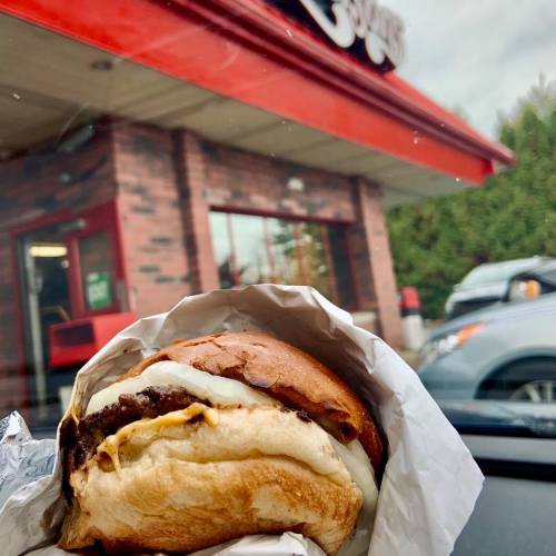 A close-up of a burger wrapped in paper, with a fast-food restaurant in the background.