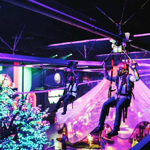 Two people ziplining indoors, surrounded by colorful lights and decorations in a vibrant, festive atmosphere.