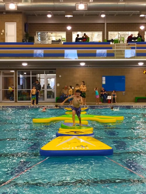 A child balances on floating mats in a swimming pool, with other kids and adults watching from the sidelines.