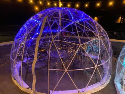 A clear geodesic dome with blue lights, set outdoors on a patio, surrounded by string lights at night.