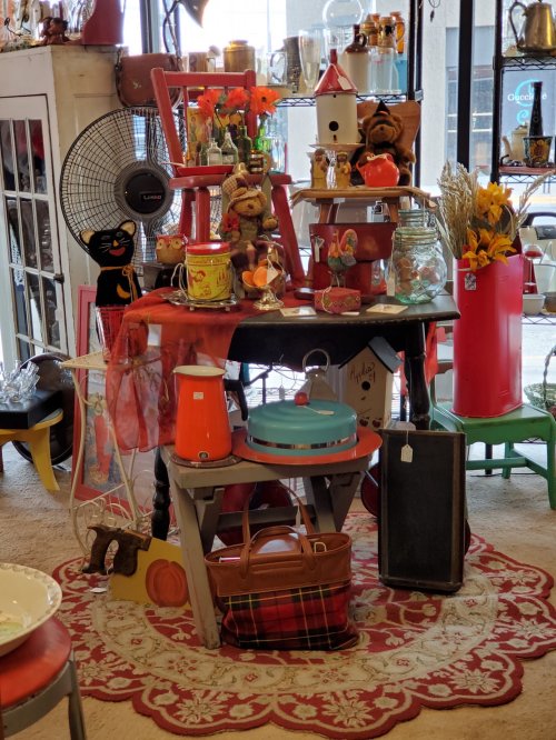 A colorful display of vintage items, including chairs, decor, and kitchenware, arranged on a table with a patterned rug.