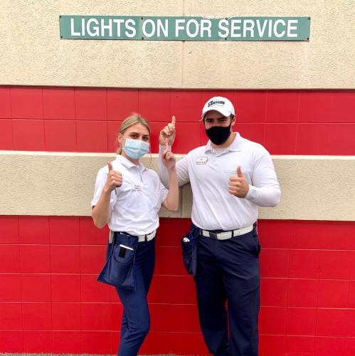 Two employees in uniforms and masks stand in front of a "Lights On for Service" sign, giving thumbs up.
