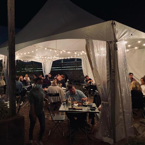 A lively outdoor dining area under a tent, filled with people enjoying a meal, illuminated by string lights at night.