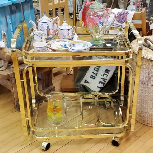 A vintage gold bar cart with glass shelves, featuring teapots, a vase, and a pitcher, set in a cozy shop.