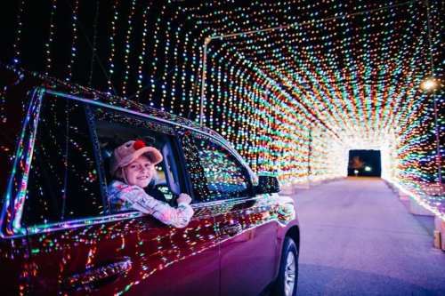 A child in a car wearing a hat smiles from the window, surrounded by a colorful tunnel of holiday lights.