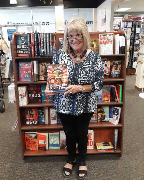 A woman holds a book titled "Killing the Rising Sun" in a bookstore, surrounded by shelves of various books.