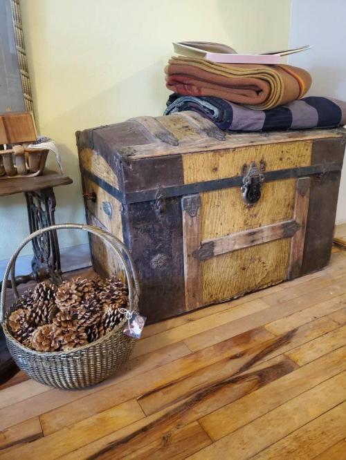 A vintage wooden trunk with a metal latch, topped with folded blankets, beside a woven basket of pinecones on a wooden floor.