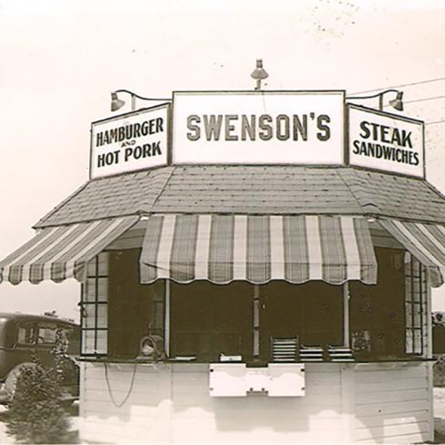 Black and white photo of a vintage food stand with a striped awning, featuring signs for hamburgers and steak sandwiches.