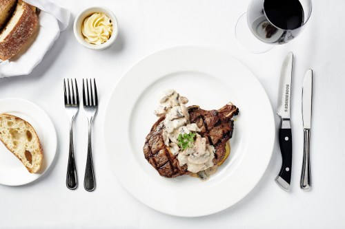 A plated steak topped with creamy sauce, accompanied by bread, butter, and a glass of red wine on a white tablecloth.