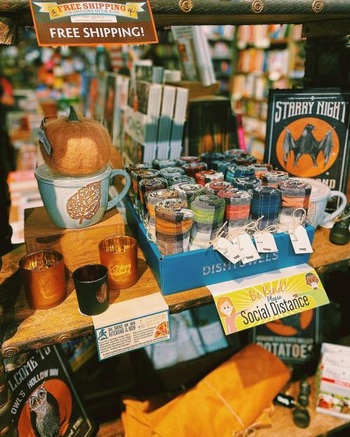 A cozy display of candles, mugs, and colorful dish towels in a bookstore, with signs for free shipping and social distancing.