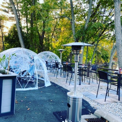 Outdoor dining area featuring clear igloo-like structures, tables, and a patio heater surrounded by trees.