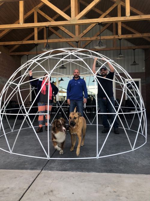 Four people and two dogs stand inside a large white geodesic dome structure in a spacious outdoor area.