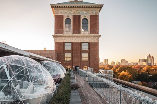 Rooftop view featuring transparent igloos, a brick building, and a city skyline at sunset.