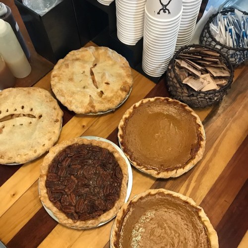 A variety of pies on a wooden table, including pecan, pumpkin, and a decorative crust pie, with cups and condiments nearby.