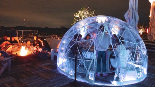 A cozy igloo-shaped structure with people dining inside, surrounded by a fire pit and a nighttime cityscape.