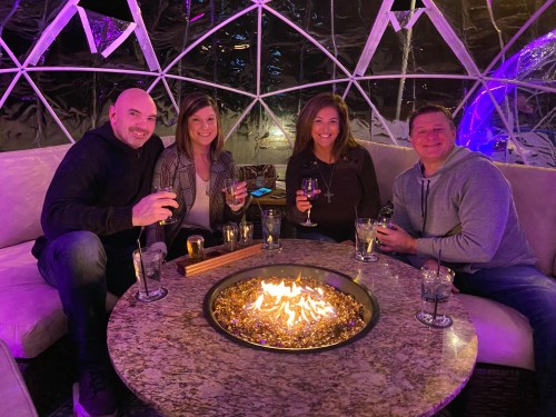 A group of four friends enjoying drinks around a cozy fire pit inside a stylish igloo-like structure.